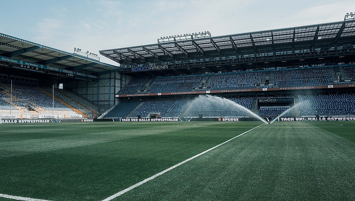 Das Stadion der Schüco-Arena mit leeren Rängen und einem laufenden Rasensprenger vor der Stadionöffnung.
