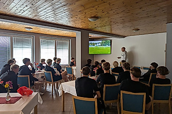 Die U21-Mannschaft sitzt in einem Raum und schaut auf einen Fernsehr, wo Cheftrainer Sebastian König eine Videoanalyse durchführt.