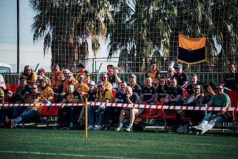 Fans von Dynamo Dresden sitzen im Trainingslager am Spielfeldrand.