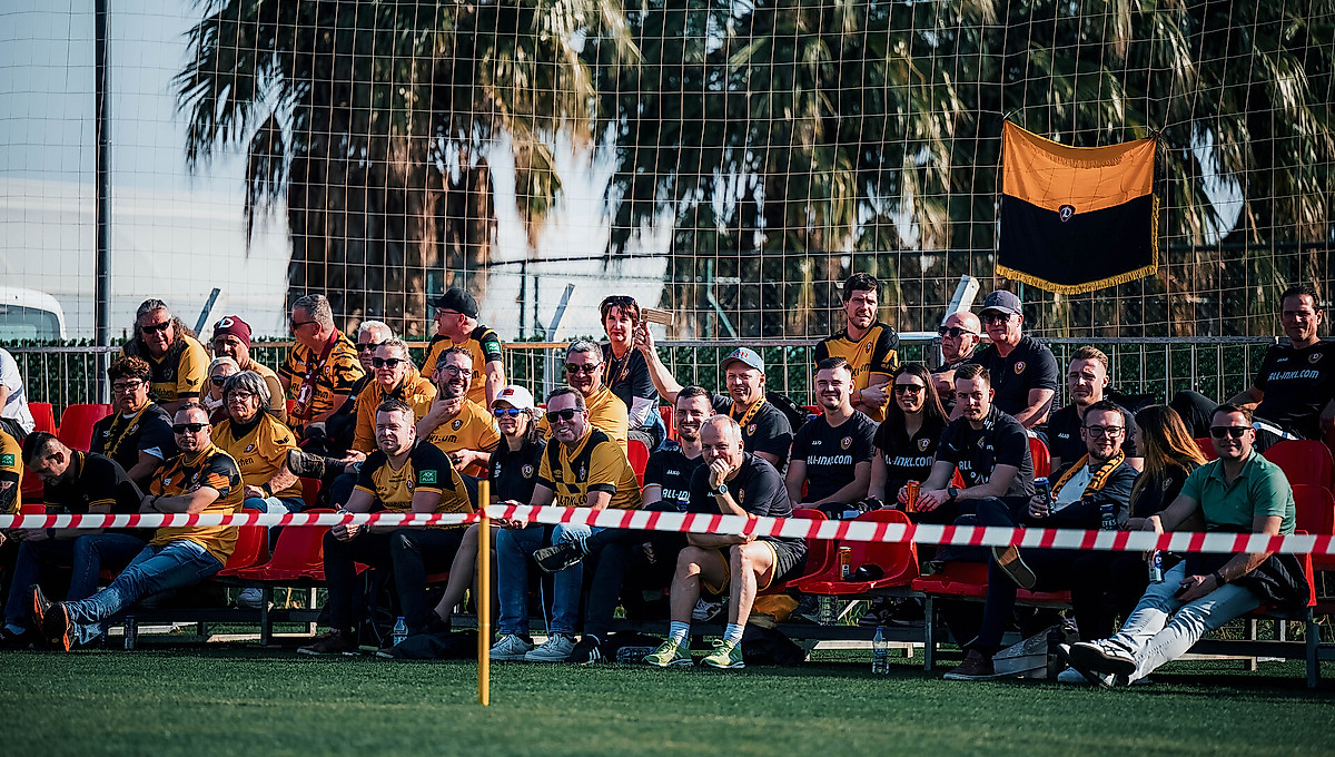 Fans von Dynamo Dresden sitzen im Trainingslager am Spielfeldrand.
